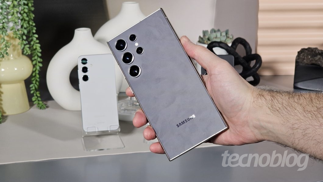 Mão segurando smartphone, ao fundo outro smartphone em um suporte de acrílico sobre uma mesa