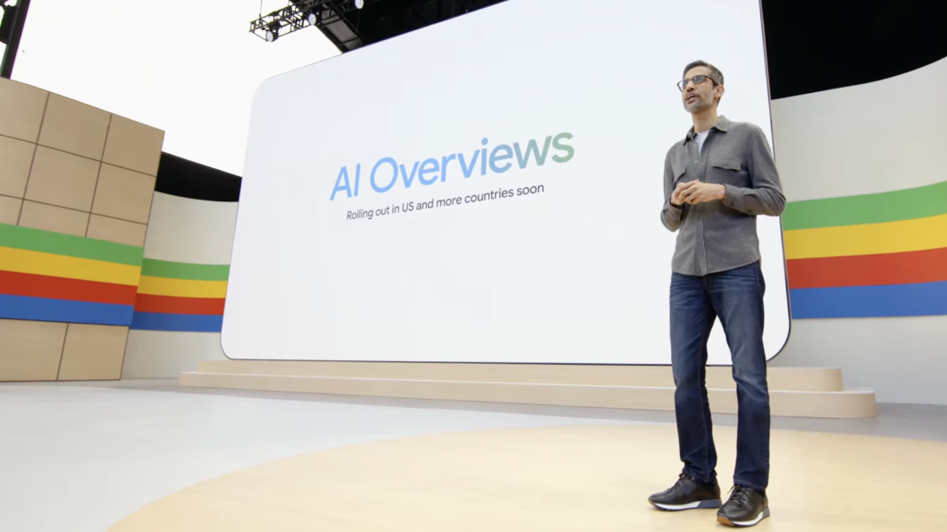 Homem sobre palco. Ao fundo, um telão exibe os escritos "AI Overviews"