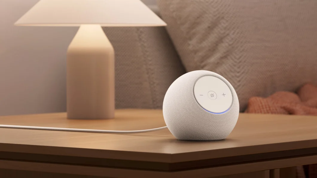 Um alto-falante inteligente Echo Dot Max branco, em formato esférico, está sobre uma mesa de madeira clara ao lado de uma luminária acesa. O dispositivo tem botões “+”, “–” e de microfone na parte superior, com um anel de LED azul iluminado. Ao fundo, há travesseiros e uma manta em tons neutros, compondo um ambiente de quarto aconchegante.