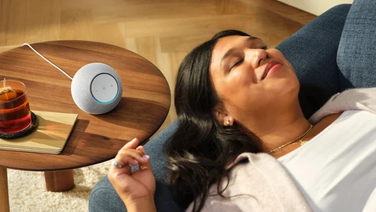 Mulher deitada em um sofá, de olhos fechados e expressão relaxada. Ao lado dela, sobre uma mesa de madeira, há um alto-falante inteligente pequeno e redondo, com luz azul acesa na parte superior, indicando ativação da assistente de voz. Há também um copo de chá em um pires. A cena transmite conforto e tranquilidade em um ambiente doméstico iluminado.