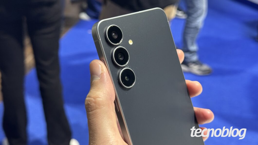 Uma mão segura um smartphone Galaxy S25 FE cinza e preto. O aparelho é exibido de perfil, mostrando o acabamento metálico e o módulo de câmera com três lentes circulares alinhadas verticalmente no canto superior. O flash está localizado ao lado das lentes. O fundo é desfocado, mostrando pessoas e um piso azul. No canto inferior direito, o logotipo "tecnoblog" está em branco.