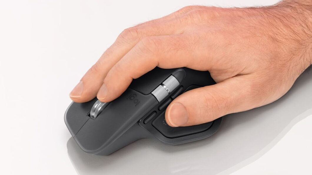 Uma mão segura um mouse sem fio preto da Logitech, posicionado sobre uma superfície branca. O polegar repousa sobre uma área lateral com botões e uma roda de rolagem metálica, enquanto o indicador e o médio estão sobre os botões principais e a roda de rolagem superior. O design é ergonômico e moderno.