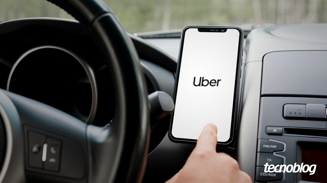 Imgem mostra uma pessoa ao volante, teclando sobre um celular preso ao lado do volante. O aparelho está abrindo o aplicativo Uber. Na parte inferior direita, o logotipo do "tecnoblog" é visível.