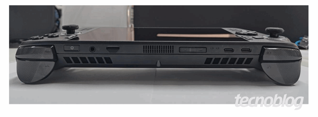 Foto em close-up, vista lateral, de um console de videogame portátil de cor preta, mostrando a parte superior do dispositivo. Na parte central, há orifícios de ventilação, e nas laterais, as pegadas ergonômicas. A borda superior do console exibe, da esquerda para a direita: um botão de liga/desliga, um conector P2 para fone de ouvido, um slot para cartão, botões de volume, e portas USB-C. O logo do Tecnoblog aparece no canto inferior direito.