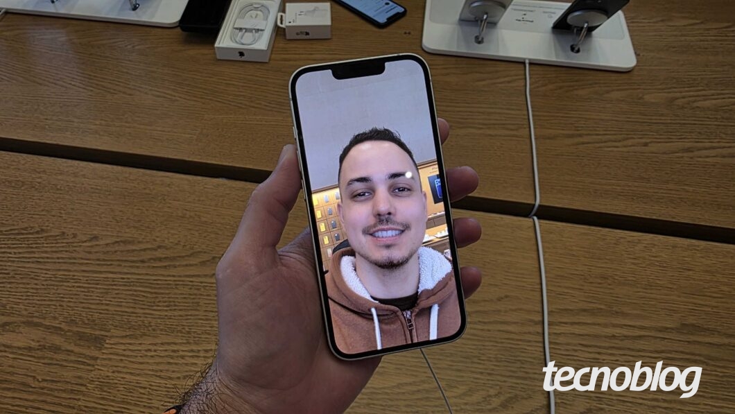 Fotografia colorida mostra um iPhone 16e sendo segurado em uma mão. Ele está com a câmera frontal aberta.