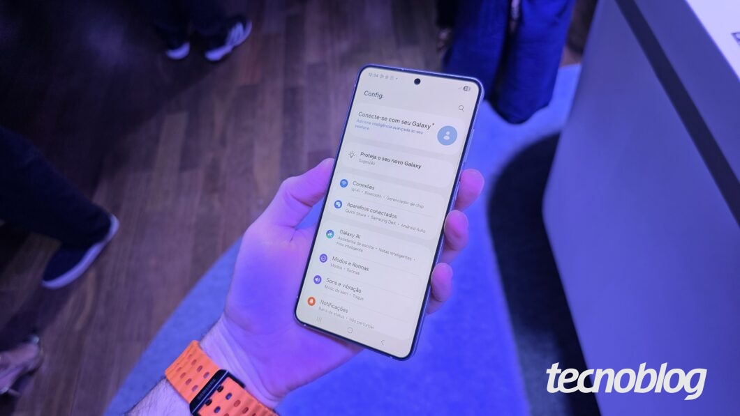 Uma mão segura um smartphone Samsung Galaxy S25 Edge com a tela ligada, exibindo o menu de "Configurações". O topo da tela mostra "Config." e "Conecte-se ao seu Galaxy", seguido de "Proteja o seu novo Galaxy". Abaixo, uma lista de opções como "Conexões", "Aparelhos conectados", "Galaxy AI", "Modos e rotinas" e "Sons e vibração" é visível. Na parte inferior direita, o logotipo do "tecnoblog" é visível.