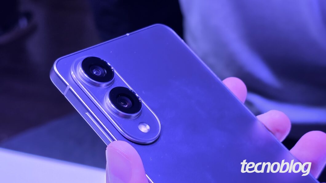 Imagem colorida mostra um smartphone Samsung Galaxy S25 Edge sendo segurado em uma mão, com a traseira para cima. A foto está em close-up nas duas câmeras traseiras. Na parte inferior direita, o logotipo do "Tecnoblog" é visível.