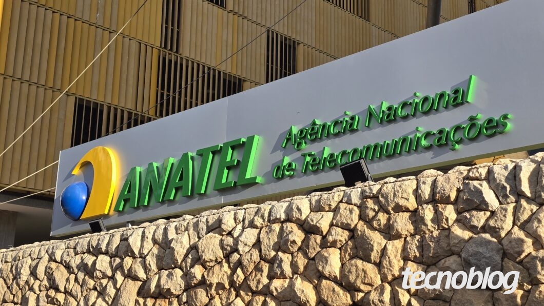 Placa da Anatel (Agência Nacional de Telecomunicações) fixada sobre um muro de pedras. A placa exibe um logotipo com uma forma curva amarela envolvendo uma esfera azul, seguido do texto "ANATEL" em letras maiúsculas verdes. Ao lado, em letras verdes menores, está escrito "Agência Nacional de Telecomunicações". Ao fundo, parte da fachada do prédio com estruturas verticais amarelas.