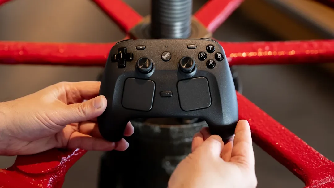  Duas mãos seguram firmemente um controle de videogame preto com o logotipo do Steam no centro superior. O controle tem um design moderno com dois analógicos, um direcional digital em formato de cruz à esquerda e os botões coloridos "A", "B", "X" e "Y" à direita. Dois grandes botões retangulares, que parecem ser touchpads, estão posicionados abaixo dos analógicos.