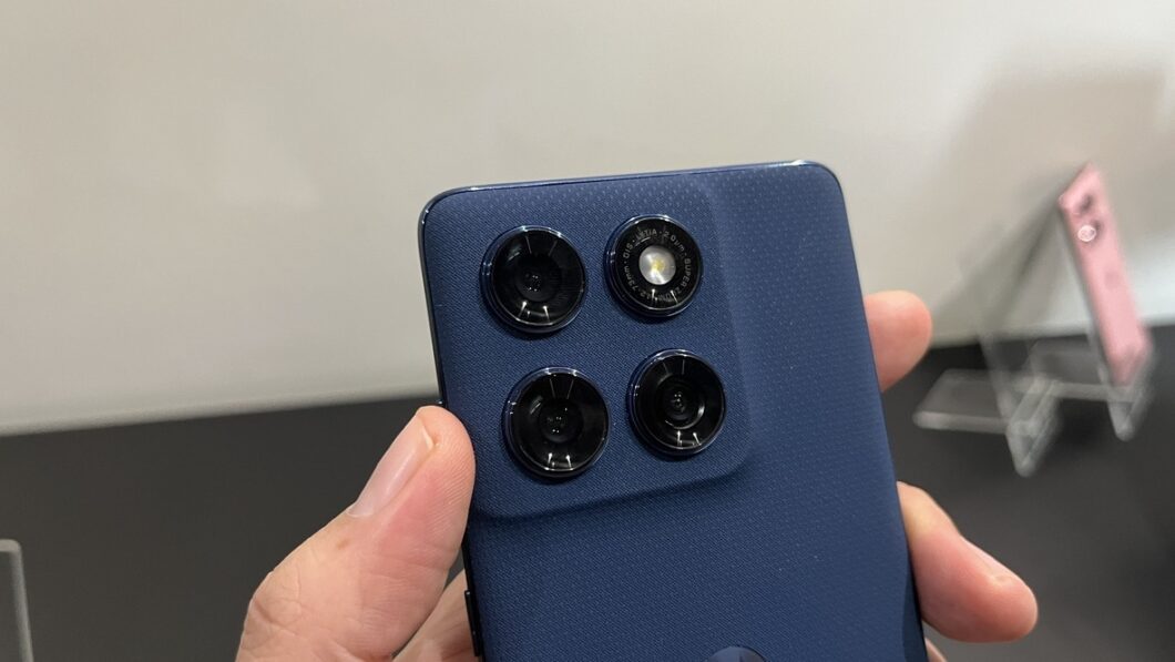 A imagem mostra a parte traseira do Motorola Edge 60, com foco na área das câmeras. O dispositivo possui um módulo de câmeras quadrado, com três lentes circulares e um flash LED. O aparelho é de cor azul escuro e está sendo segurado com uma mão. Ao fundo, é possível ver um suporte transparente com outros aparelhos.