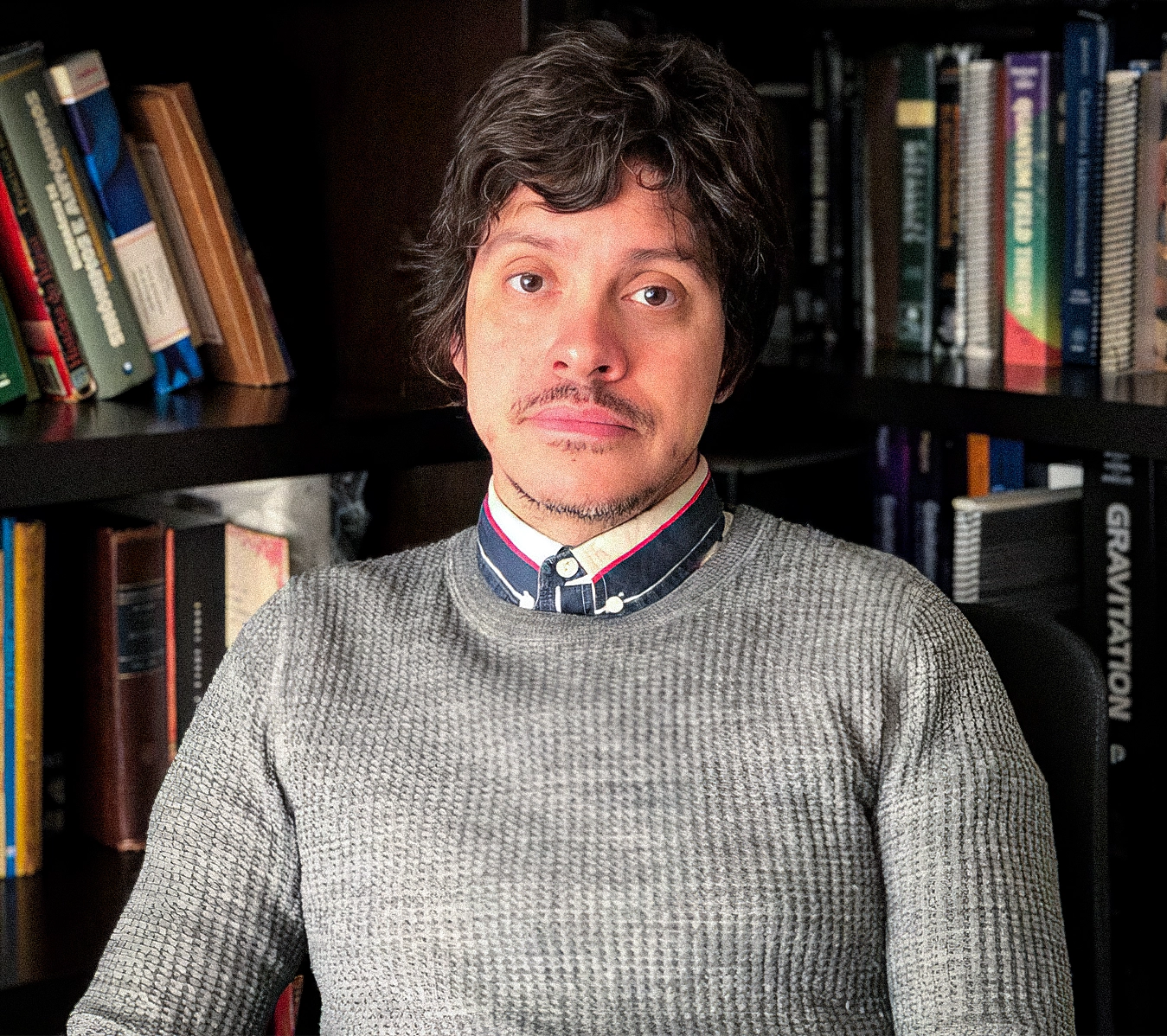 A young man sits in front of a bookshelf.