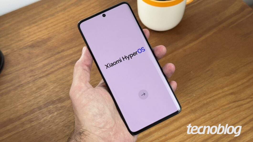 Fotografia colorida do celular Poco X7 da Xiaomi, de cor preta e amarela, fotografado de frente. Ele é segurado em uma mão e a tela exibe os escritos “Xiaomi HyperOS”.