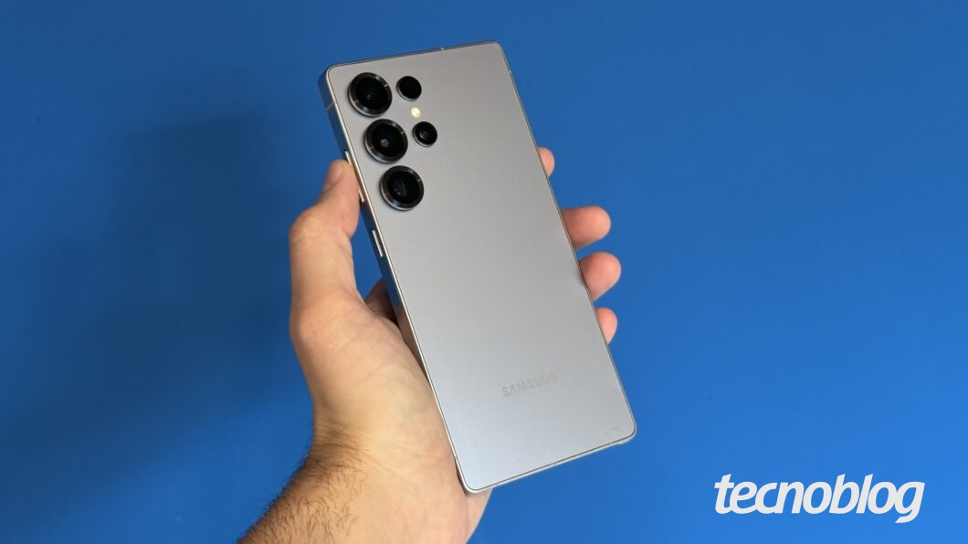 Uma mão segura um smartphone Samsung de cor prateada contra um fundo azul. O aparelho possui um design premium com cantos retos e uma configuração de câmera traseira composta por quatro lentes dispostas verticalmente no canto superior esquerdo. O logo da Samsung está gravado na parte inferior traseira do dispositivo.