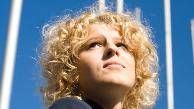 Young adult with curly blonde hair, looking upwards with a bright sky in background