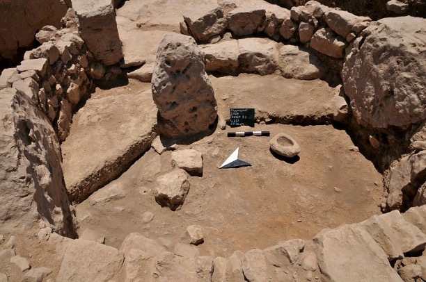 An oblong stone standing upright in the middle of circular stone walled enclosure with no roof. A mortar sits to the right of the central stone, and archeological markers have been placed at the base of the stone.