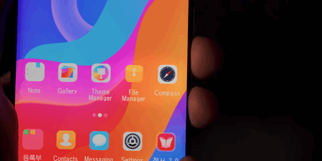 Close-up da tela inicial de um smartphone norte-coreano (Samtaesung ou similar) exibindo uma grade de ícones de aplicativos sobre um papel de parede vibrante e colorido (azul, rosa, laranja). Ícones visíveis incluem 'Note', 'Gallery' (Galeria), 'Theme Manager' (Gerenciador de Temas), 'File Manager' (Gerenciador de Arquivos) e 'Compass' (Bússola). A parte inferior da tela mostra ícones para 'Contacts' (Contatos), 'Messaging' (Mensagens) e 'Settings' (Configurações).