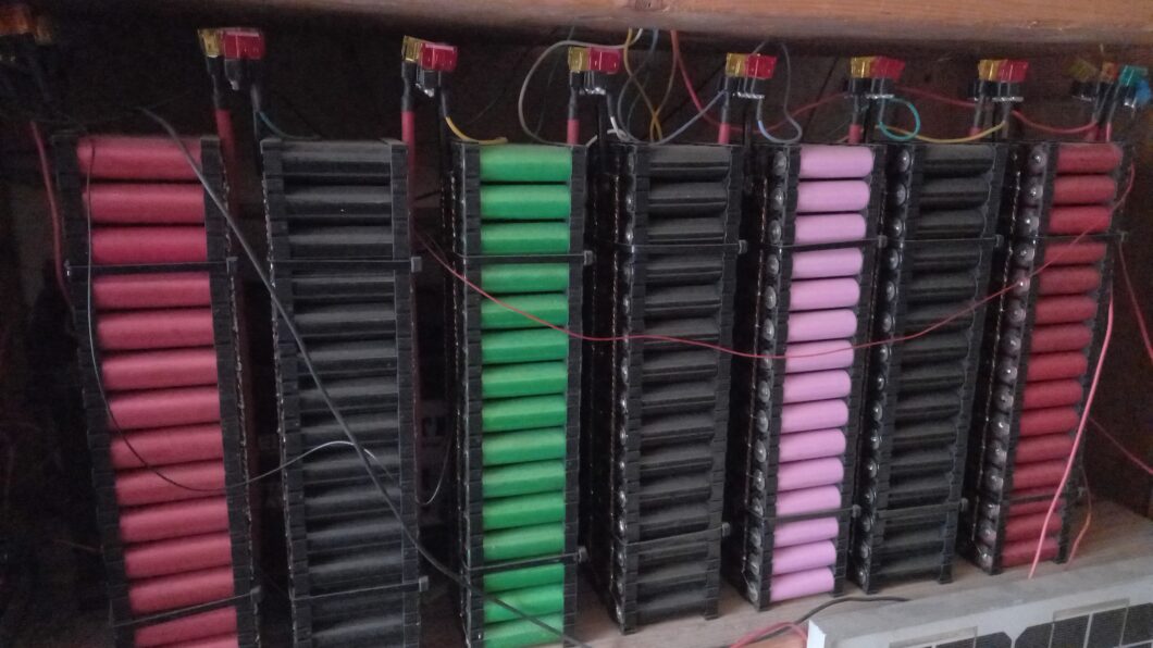 Muralha de módulos de armazenamento de energia construídos com pilhas verticais de células de bateria de íon de lítio (18650). As células são dispostas em racks pretos, com algumas colunas em vermelho, verde e rosa. Cabos e fusíveis de segurança vermelhos estão conectados ao topo das colunas, sugerindo um sistema de bateria de alta capacidade, feito sob medida, para uso em casa ou em sistemas solares off-grid.