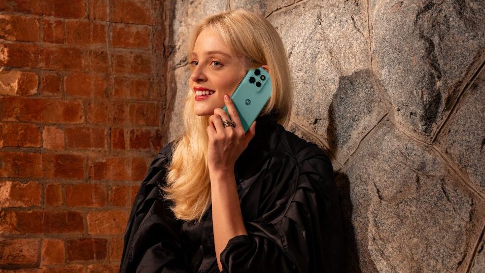 Foto de uma mulher loira de pele clara, sorrindo e olhando para o lado, como se estivesse conversando ao telefone. Ela segura um smartphone Motorola Edge 60 Neo de cor verde-água encostado à sua orelha.