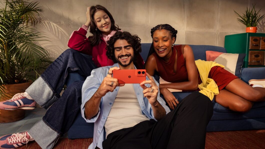 Foto de três jovens adultos sorrindo, sentados no chão e em um sofá, em um ambiente decorado com plantas e almofadas. O homem no centro, com cabelo cacheado escuro, segura um smartphone Motorola Edge 60 Neo vermelho-coral na horizontal, como se estivesse filmando ou tirando uma foto com a câmera traseira