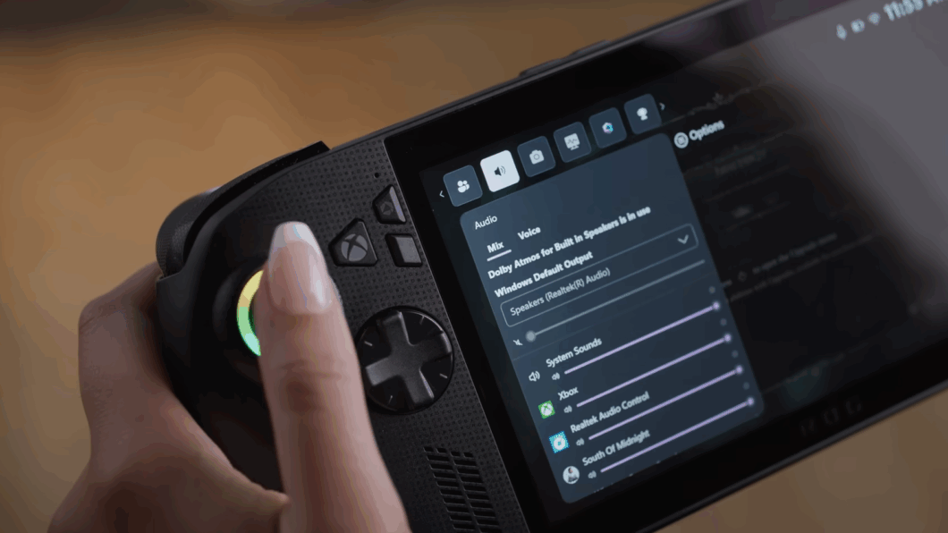 Close-up de uma mão segurando um console de videogame portátil preto (tipo ROG Ally). Um dedo pressiona o botão de função superior ao lado de um joystick com um anel de luz verde e amarela.