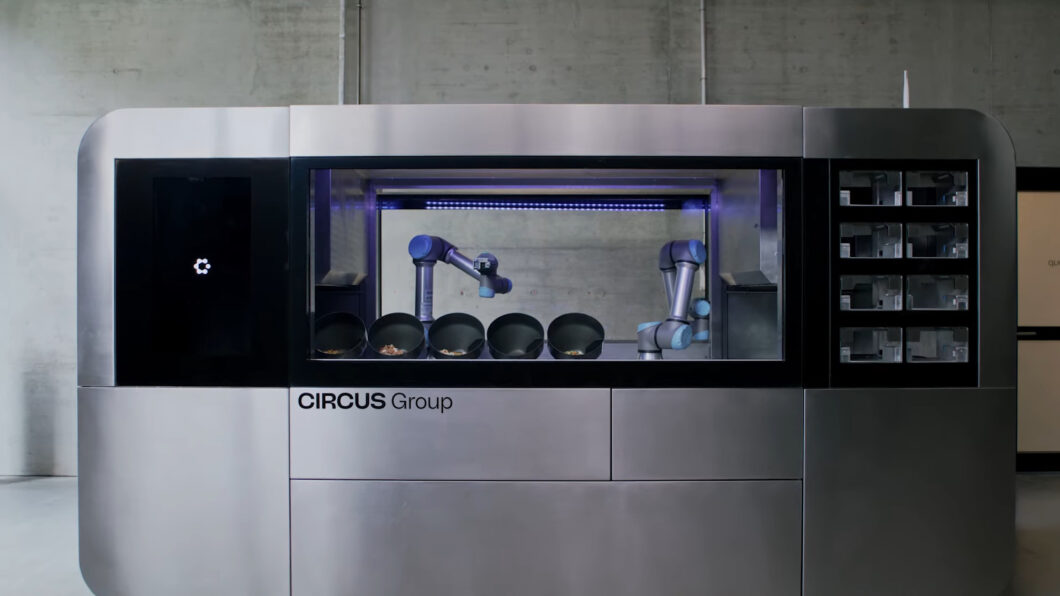 Máquina de cozinha industrial automatizada, com estrutura metálica grande e retangular. No centro, braços robóticos manipulam panelas internas atrás de um vidro. À esquerda há uma tela preta, e à direita, compartimentos de armazenamento. Na parte frontal, lê-se “CIRCUS Group”. A cena sugere preparo de refeições sem intervenção humana.