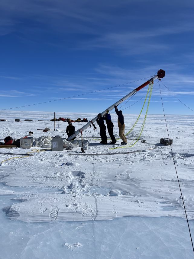 Three people standing beneath a long, silver column, raising it into a vertical position. A fourth persons kneels at the instrument's base, which is surrounded by boxes. The people are standing on ice, and the sky is blue with a few wispy clouds.