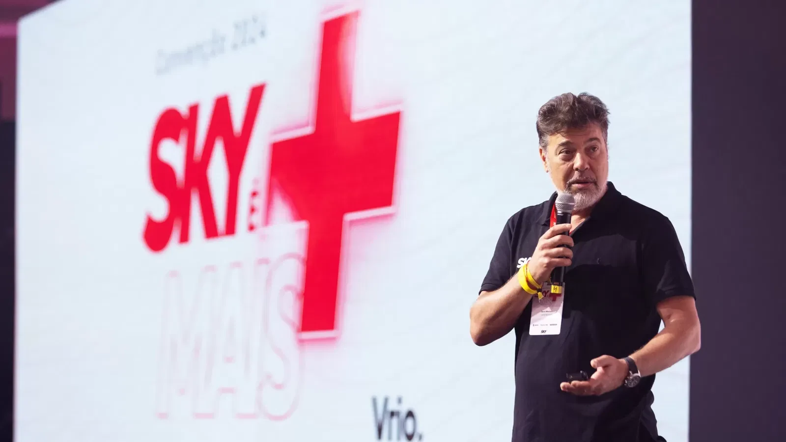 Um homem, com cabelos grisalhos e barba, usando uma camisa polo preta, fala segurando um microfone durante uma apresentação.