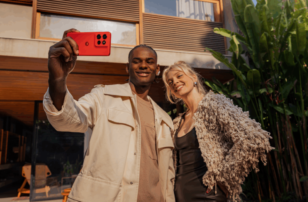 Duas pessoas posam para uma selfie ao ar livre enquanto uma delas segura um smartphone Motorola vermelho, voltado para a câmera. O aparelho exibe três lentes traseiras e o logotipo da marca. A dupla sorri em frente a uma casa moderna, com plantas tropicais ao fundo e luz natural destacando o design vibrante do celular.
