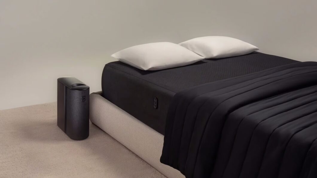 Cama com colchão e cobertor pretos em um quarto minimalista, com duas almofadas brancas sobrepostas na cabeceira. Ao lado da cama, no chão, há um aparelho alto e estreito, também preto, parte de um sistema de controle de temperatura ou monitoramento do sono. O ambiente é neutro, com paredes claras e carpete bege.