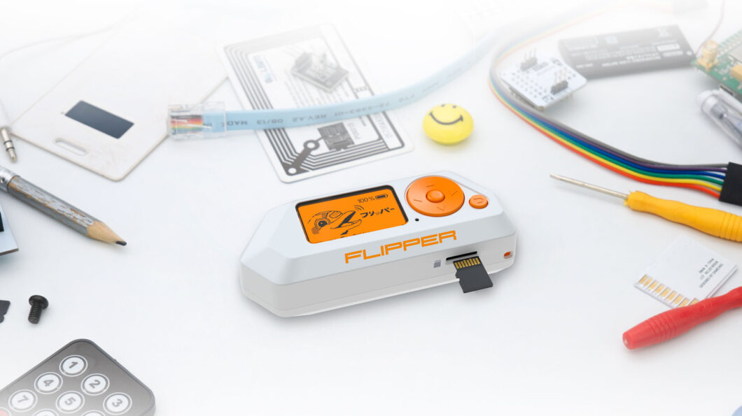 Dispositivo eletrônico branco e laranja Flipper Zero sobre mesa clara, cercado por ferramentas e componentes eletrônicos, como chaves de fenda, cabos coloridos, placas de circuito e um lápis. Na tela laranja aparece um desenho de golfinho, além do indicador de bateria “100%”. O nome “FLIPPER” está impresso na lateral, e há um cartão microSD parcialmente inserido.