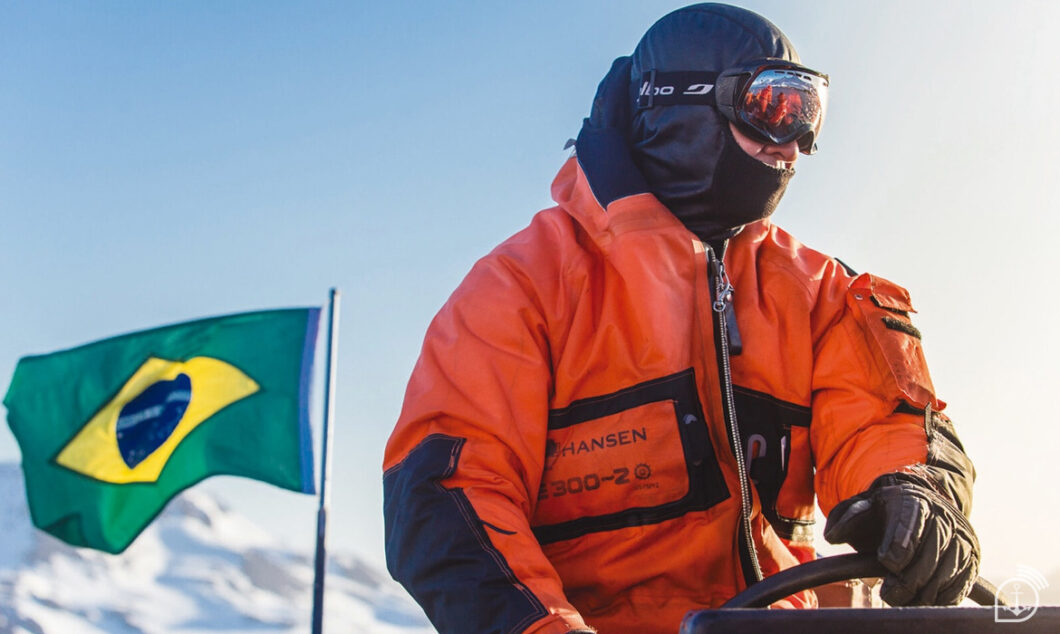 Cientista ou pesquisador brasileiro, usando um casaco de frio laranja de alta performance com capuz e balaclava preta e óculos de proteção (goggles) laranja espelhados, operando algum equipamento em um ambiente antártico. A Bandeira do Brasil tremula ao vento no canto esquerdo, em frente a montanhas nevadas e sob a luz brilhante do sol, destacando a presença do PROANTAR (Programa Antártico Brasileiro).