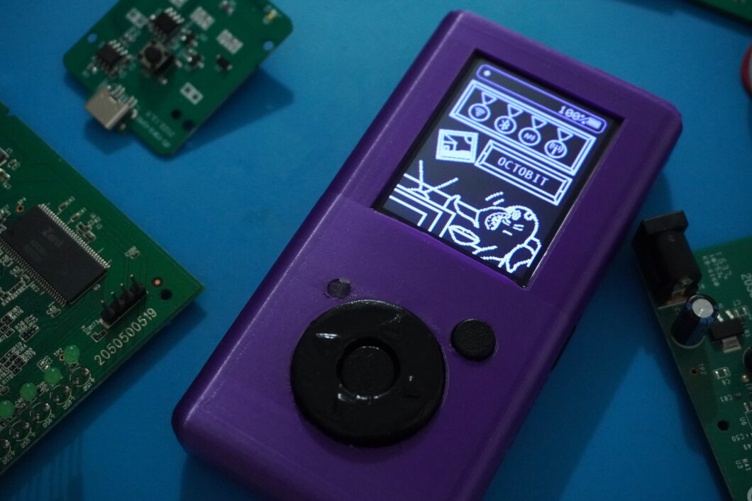 Dispositivo eletrônico roxo apoiado sobre superfície azul, cercado por placas de circuito verde. A tela exibe uma interface em preto e branco com ilustração de um polvo e o nome “OCTOBIT”, além de ícones e indicador de bateria “100%”. O aparelho tem botão direcional circular e um botão menor ao lado, com acabamento fosco e levemente gasto.