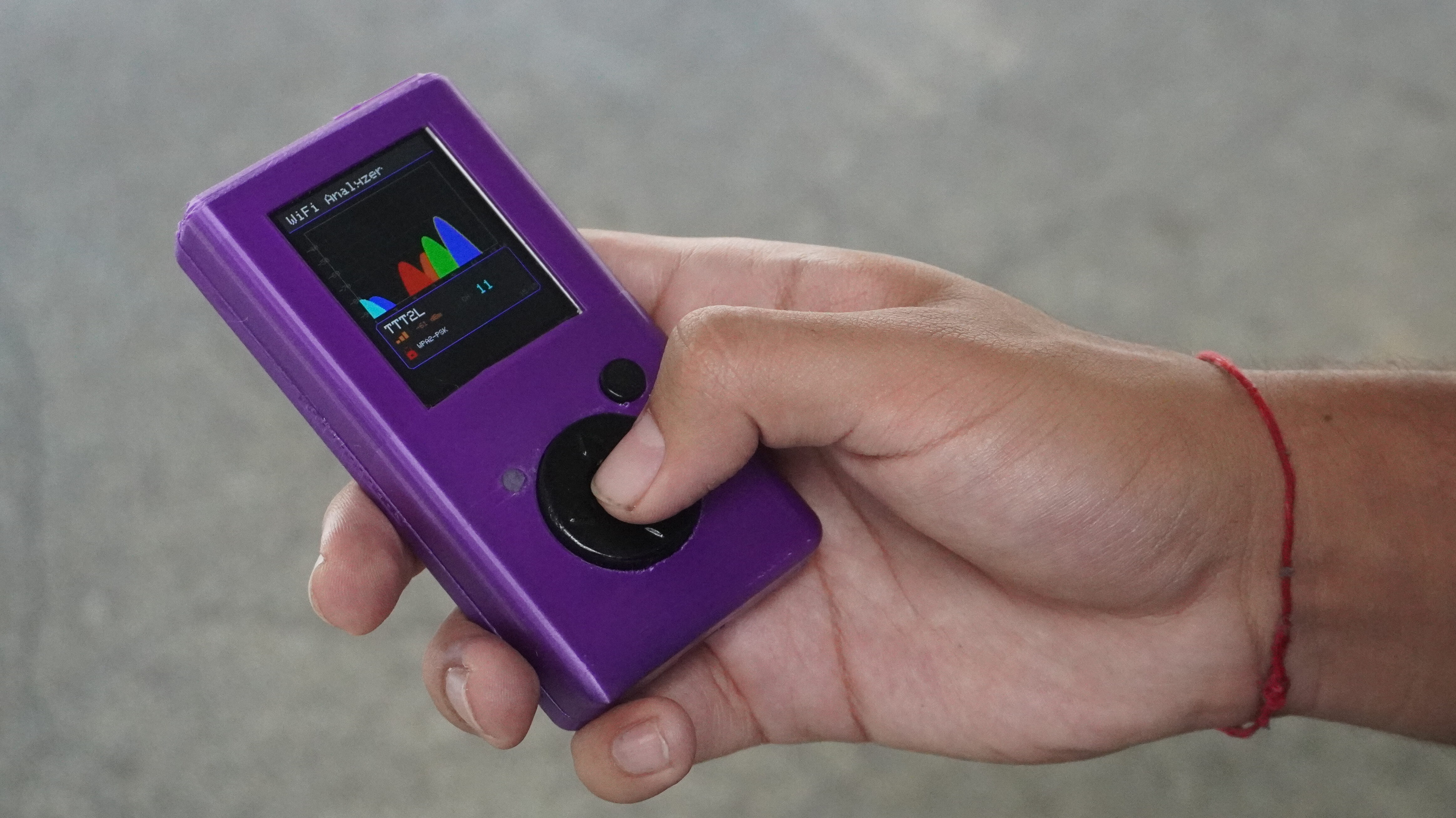 Alt text sugerido: Mão segura um dispositivo eletrônico roxo, portátil, com tela colorida e botão circular tipo joystick. Na tela aparece a interface “WiFi Analyzer”, com gráficos em formato de picos multicoloridos e indicação de rede. A pessoa usa uma pulseira vermelha no pulso, e o fundo é um piso cinza desfocado, em ambiente interno.