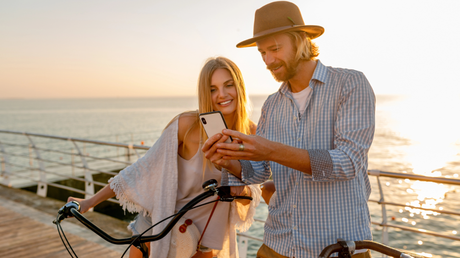 Um casal sorridente em bicicletas está parado em um píer de madeira à beira-mar, olhando juntos para a tela de um smartphone que o homem segura. O homem usa um chapéu fedora marrom e uma camisa xadrez clara, e a mulher tem cabelos longos e loiros. A cena é banhada pela luz quente do sol do pôr do sol, com o oceano ao fundo.