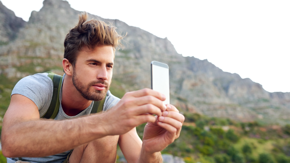 Close-up de um homem barbado em uma trilha de montanha, vestindo uma camiseta cinza e uma mochila. Ele está segurando um smartphone na horizontal com as duas mãos, olhando atentamente para a tela, possivelmente tirando uma foto ou gravando um vídeo. O fundo é composto por uma montanha rochosa e vegetação sob luz brilhante.