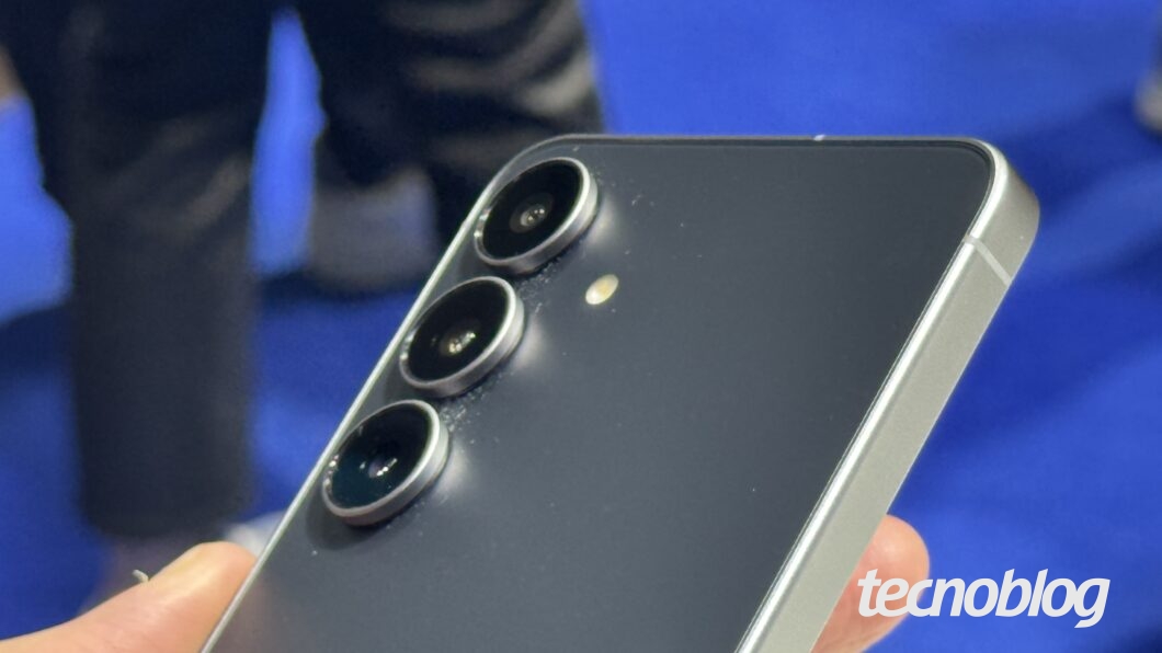 Uma mão segura um smartphone Galaxy S25 FE cinza e preto. O aparelho é exibido de perfil, mostrando o acabamento metálico e o módulo de câmera com três lentes circulares alinhadas verticalmente no canto superior. O flash está localizado ao lado das lentes. O fundo é desfocado, mostrando pessoas e um piso azul. No canto inferior direito, o logotipo "tecnoblog" está em branco.