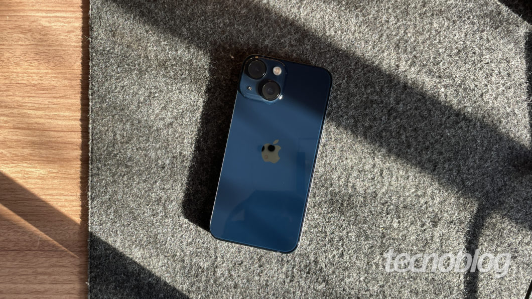 Imagem mostra um iPhone 13 Mini azul sobre um tapete felpudo. Na parte inferior direita, o logotipo do "tecnoblog" é visível. 