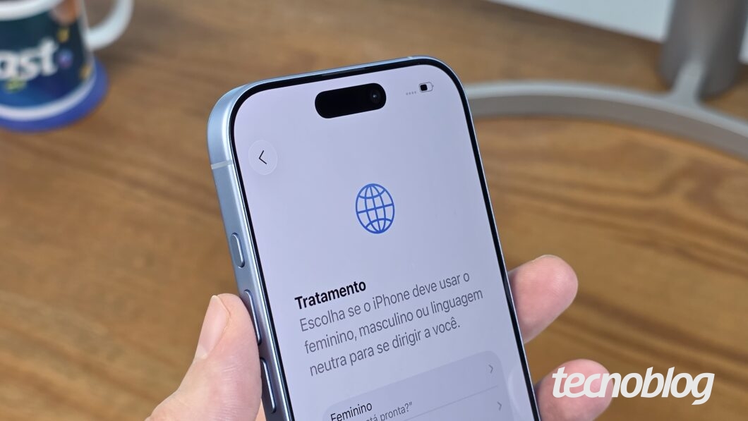 Mão segurando iPhone 17 azul-névoa, destacando a parte frontal e a lateral esquerda, enquadrando metade do aparelho, com mesa em madeira como fundo