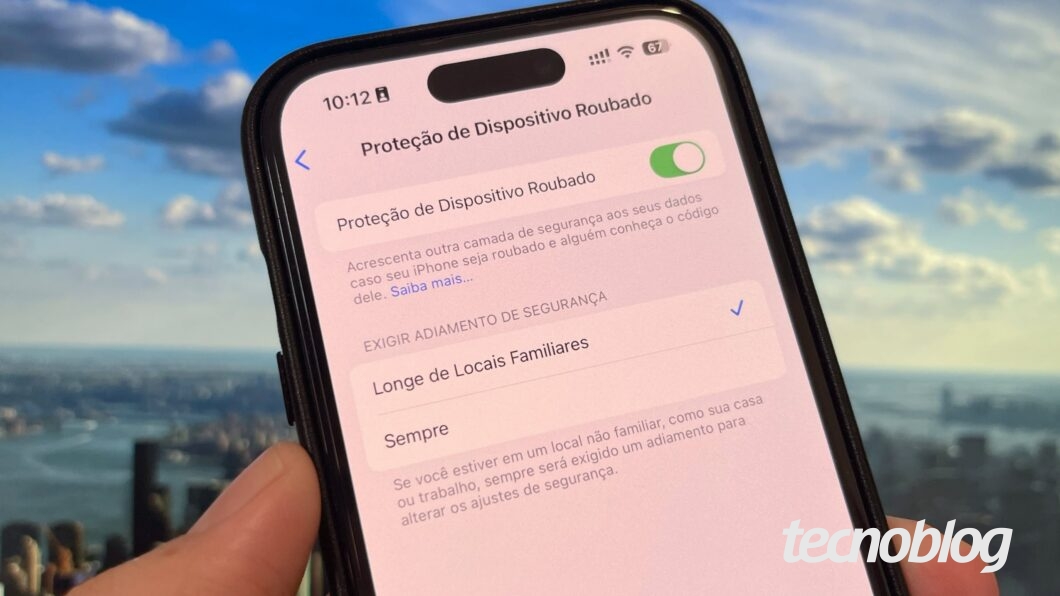 Tela de um iPhone exibindo o menu de configurações da função "Proteção de Dispositivo Roubado", com a opção ativada, indicada por um botão deslizante verde. Abaixo, há uma explicação parcial: "Acrescenta outra camada de segurança aos seus dados caso seu iPhone seja roubado e alguém conheça o código dele. Saiba mais…". Em seguida, aparecem as opções de exigência de adiamento de segurança, com "Longe de Locais Familiares" selecionada. Ao fundo, vista desfocada de uma cidade com rio, prédios e céu com nuvens.