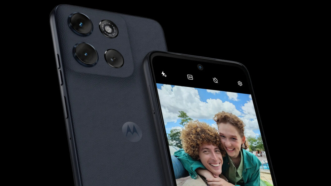 Imagem mostra um close-up de dois smartphones Motorola Moto G56 com a parte traseira em tom grafite texturizado. O aparelho em primeiro plano está ligeiramente inclinado para a direita, com a tela exibindo uma selfie de duas pessoas sorrindo para a câmera. No canto superior da tela, ícones da interface da câmera são visíveis.
