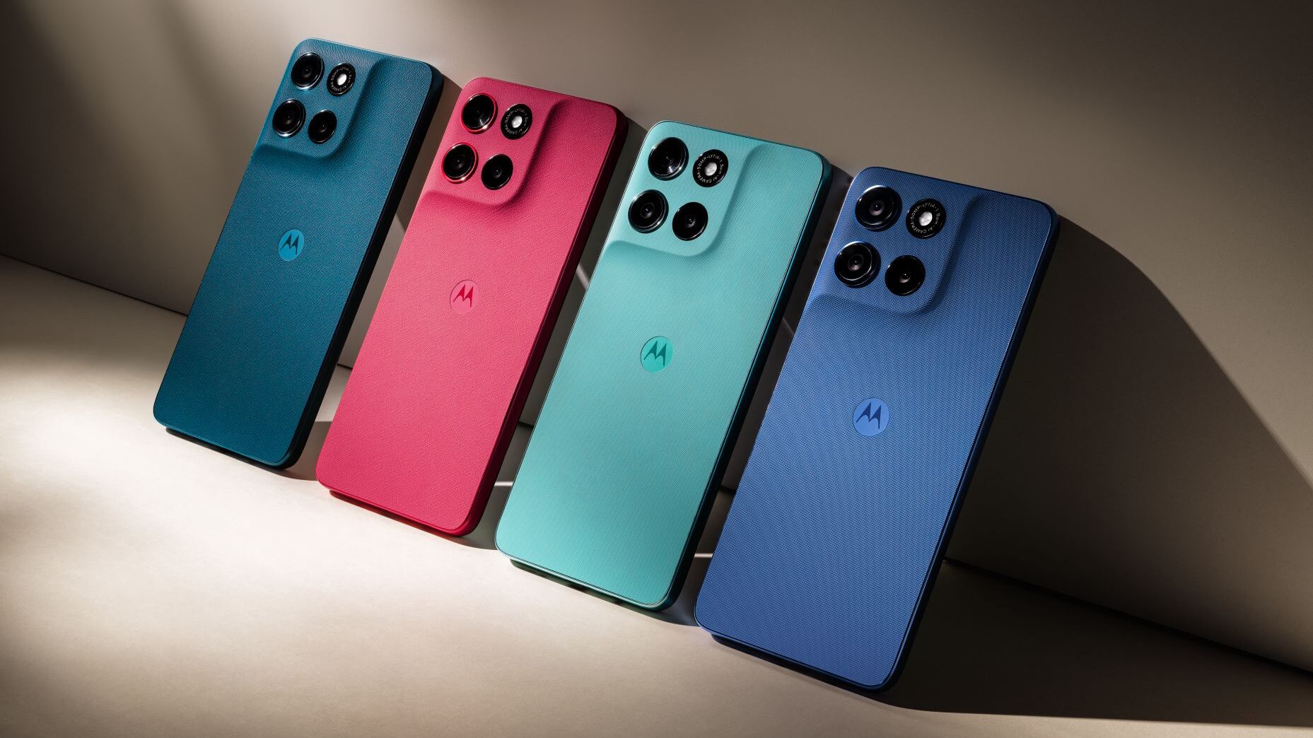 Foto de estúdio em ângulo diagonal, exibindo quatro smartphones idênticos, deitados e alinhados, em cores diferentes. Todos têm a traseira com acabamento texturizado e o logo da Motorola ao centro. Da esquerda para a direita, as cores são: azul-petróleo, rosa-choque, verde-água e azul-marinho