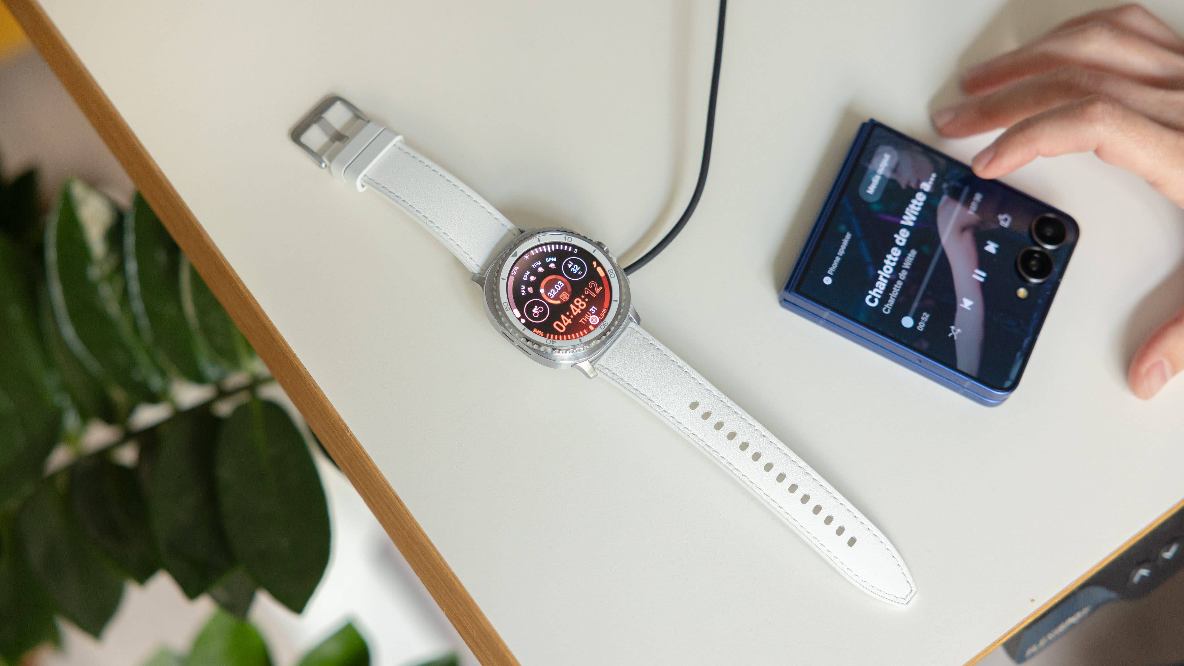 Samsung Galaxy Watch 8 Classic on a table, charging, next to a smartphone playing music.
