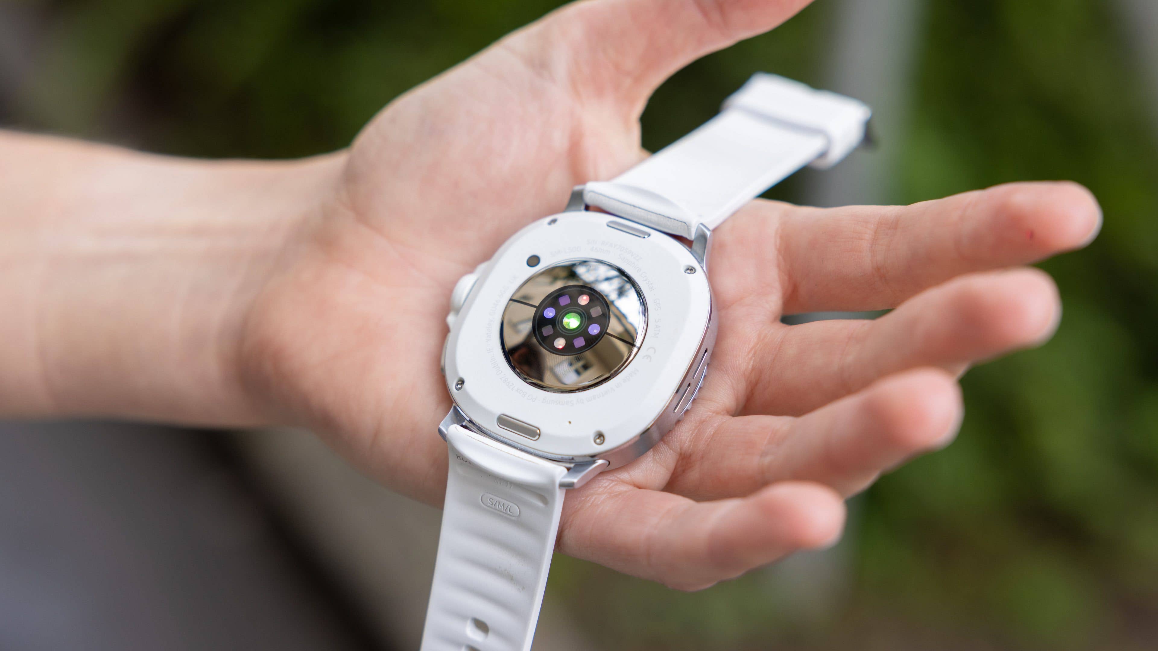 A person holding a Samsung Galaxy Watch 8 Classic showing its back with sensors and a white strap.