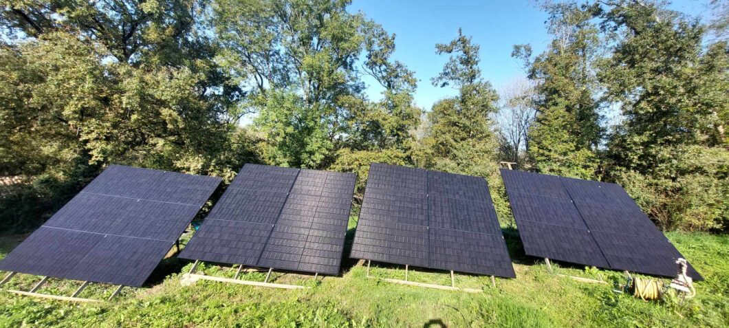 uatro painéis solares monocristalinos de cor preta, montados em estruturas de suporte inclinadas em um campo gramado. Os painéis estão dispostos em pares, voltados para o céu limpo para captação de energia. O fundo é dominado por uma densa linha de árvores verdes sob a luz do sol, sugerindo uma instalação de energia solar fora da rede ou em área rural.