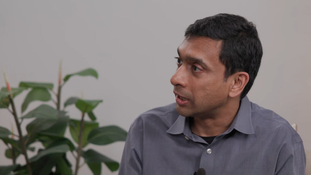 Homem de camisa social cinza aparece em um ambiente interno, de perfil, olhando para a direita. Ao fundo, há uma parede clara e uma planta com folhas verdes e brotos avermelhados.