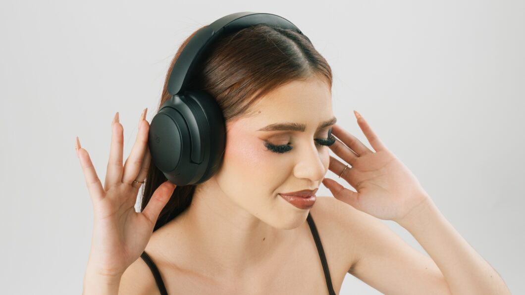 etrato de close-up de uma jovem mulher com maquiagem marcante, usando fones de ouvido QCY H3S pretos estilo over-ear, sobre um fundo branco. Ela segura as laterais dos fones com as mãos, sugerindo imersão no áudio ou ajuste do dispositivo.