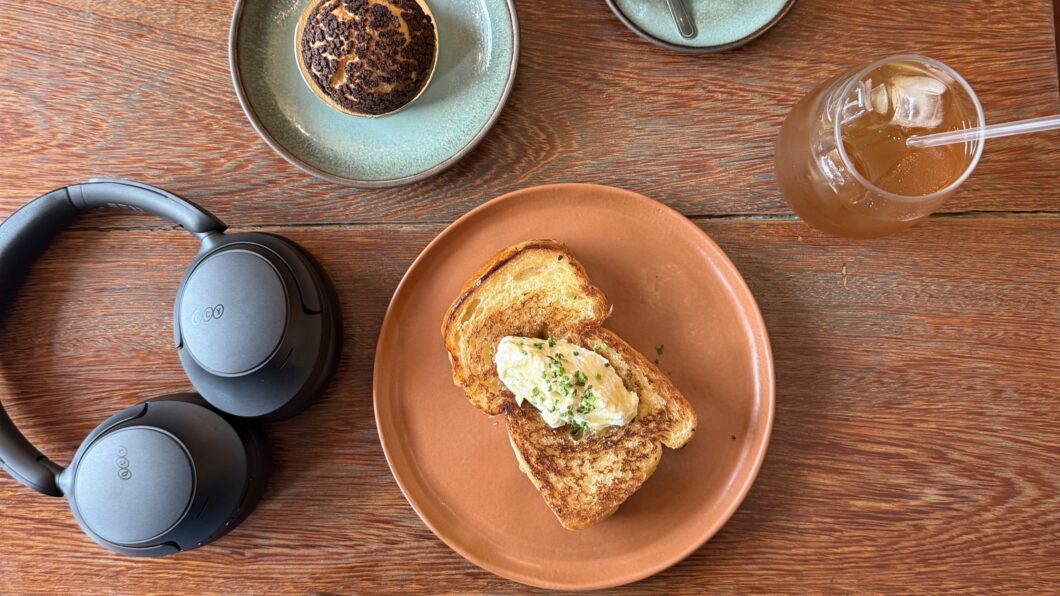Vista de cima (flat lay) de uma mesa de madeira rústica, exibindo os fones de ouvido QCY H3S pretos ao lado de um prato com uma torrada com manteiga e temperos, um pastel ou doce, e um copo de chá gelado. A imagem sugere a durabilidade da bateria para uso casual e em cafés.