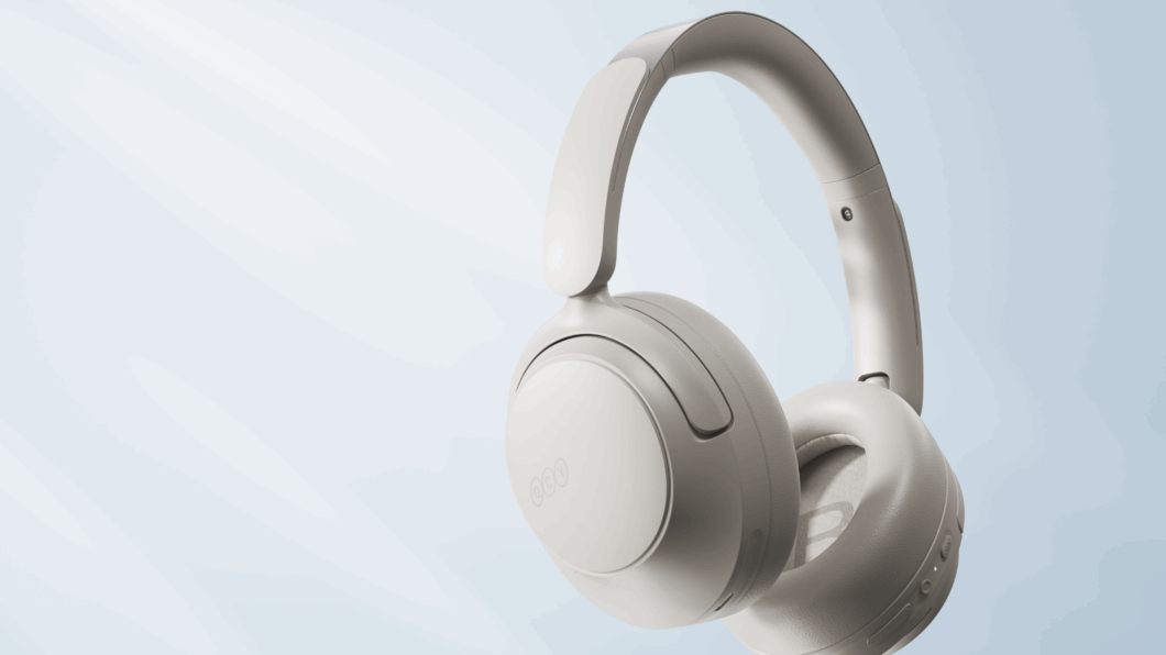 Close-up de estúdio dos fones de ouvido QCY H3S brancos/creme estilo over-ear, suspensos em um fundo cinza e azul claro, com iluminação suave. O foco está nos detalhes da concha auditiva e da alça, enfatizando a cor e o acabamento do fone.