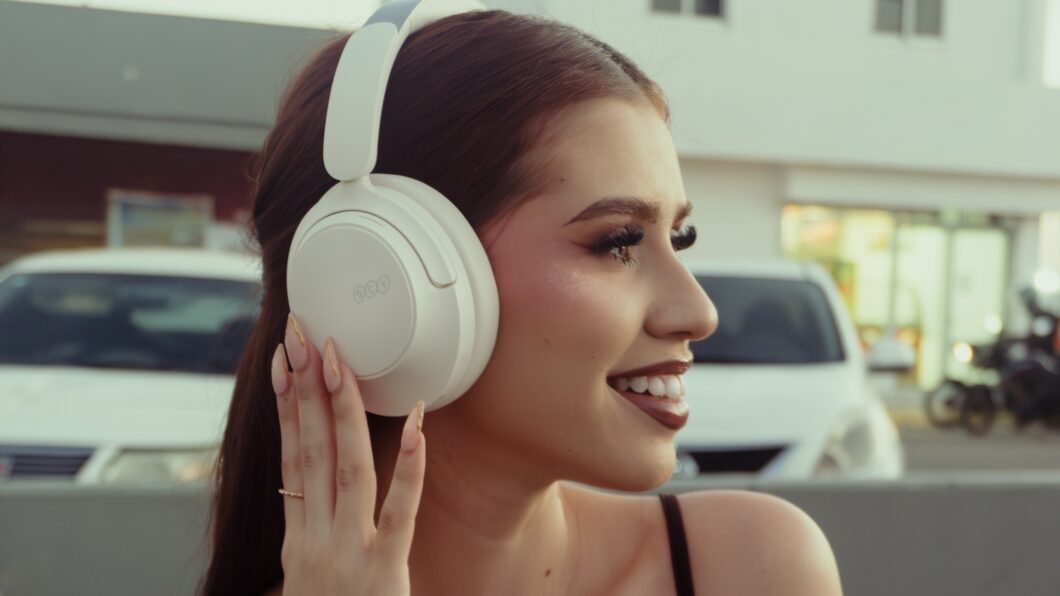 Jovem mulher sorrindo, usando fones de ouvido QCY H3S brancos em um ambiente externo e urbano, com carros brancos e motocicletas desfocados ao fundo. Ela toca o fone de ouvido direito, destacando o uso dos fones de ouvido com cancelamento de ruído (ANC) para focar em ambientes movimentados.
