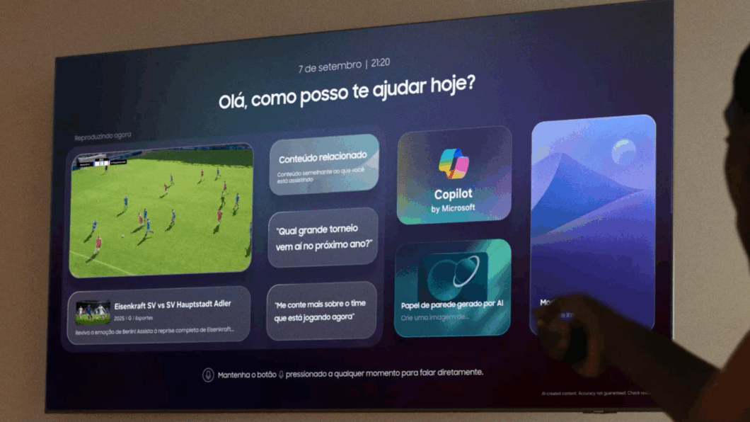 Tela de TV com interface de assistente virtual. No topo, aparecem a data e o horário “7 de setembro | 21:20” e a frase “Olá, como posso te ajudar hoje?”. À esquerda, um jogo de futebol é exibido com o texto “Reproduzindo agora”. Há cartões com sugestões como “Conteúdo relacionado”, “Qual grande torneio vem aí no próximo ano?” e “Me conte mais sobre o time que está jogando agora”. Também aparecem os ícones “Copilot by Microsoft” e “Papel de parede gerado por AI”. Uma pessoa aponta um controle remoto para a TV.